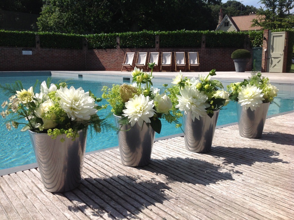 Four tall grey vessels filled with white flowers and green foliage beside a swimming pool