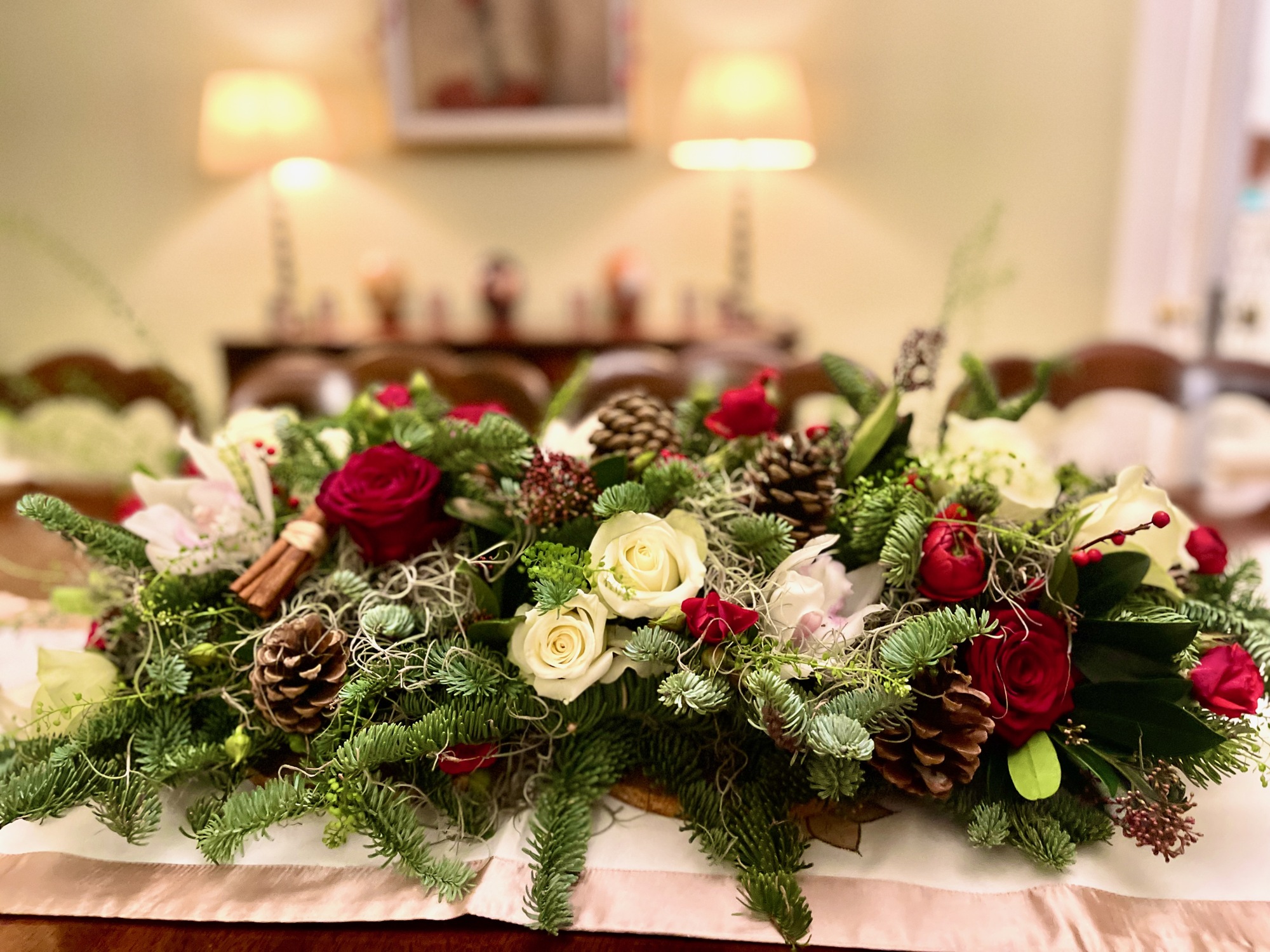 Christmas table centre with cream and red roses, pine cones and seasonal foliage, with two softly blurred lamps in the background