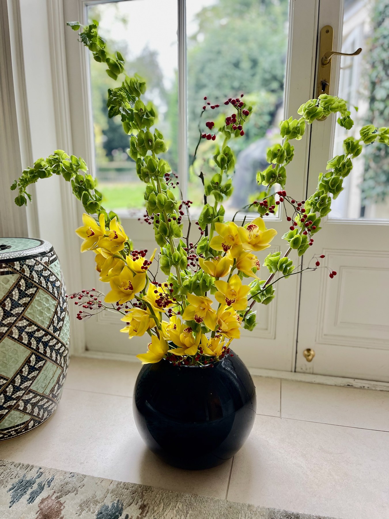 Yellow orchids, green bells of Ireland and red berries in a black vase beside a window
