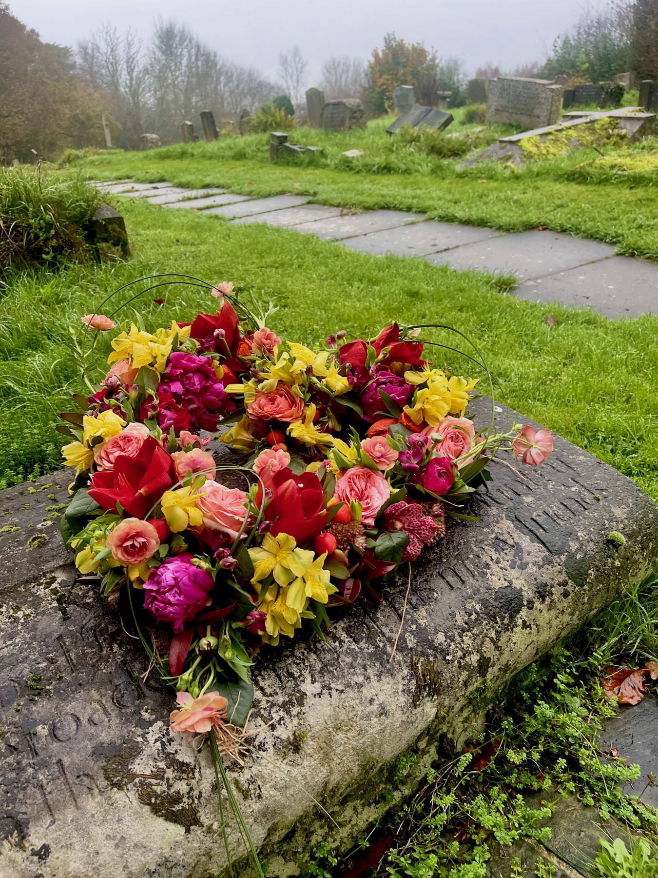 A colourful floral tribute with red, pink and yellow flowers placed on a memorial stone in a garden setting