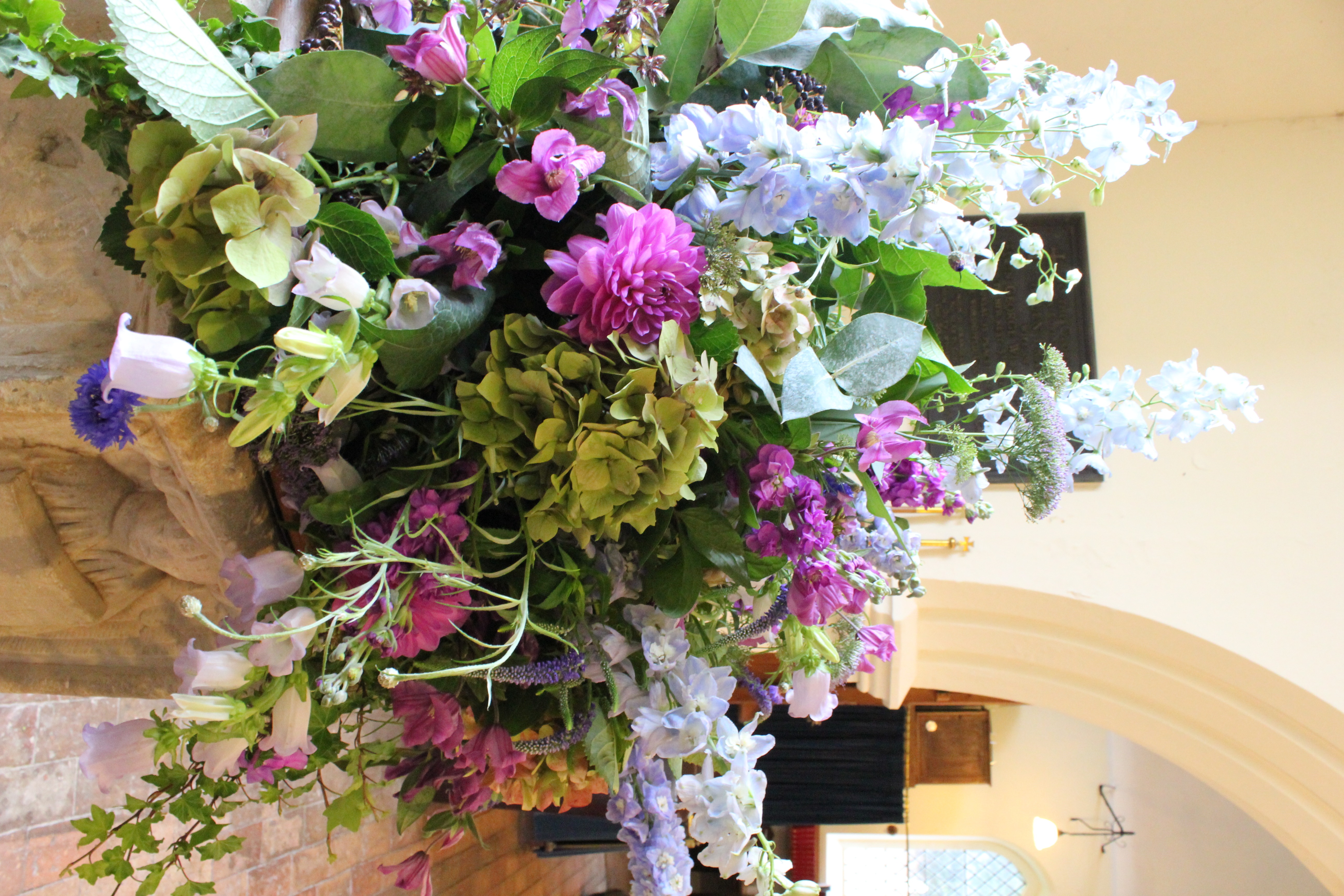 Delphiniums, purple flowers, green hydrangeas and trailing foliage arranged around a carved stone church font
