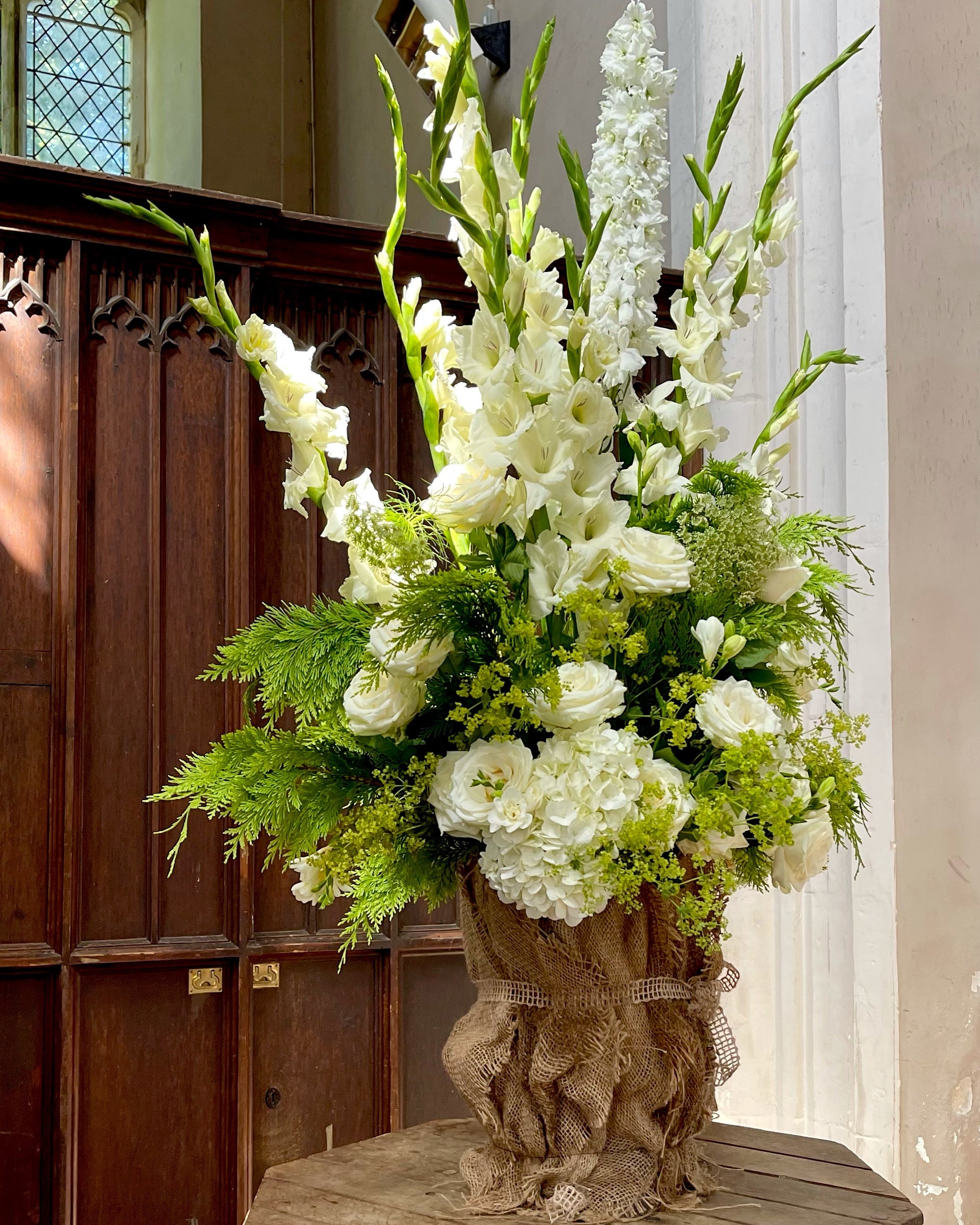 A generous white and green arrangement of gladioli, roses, hydrangea and foliage on a church font