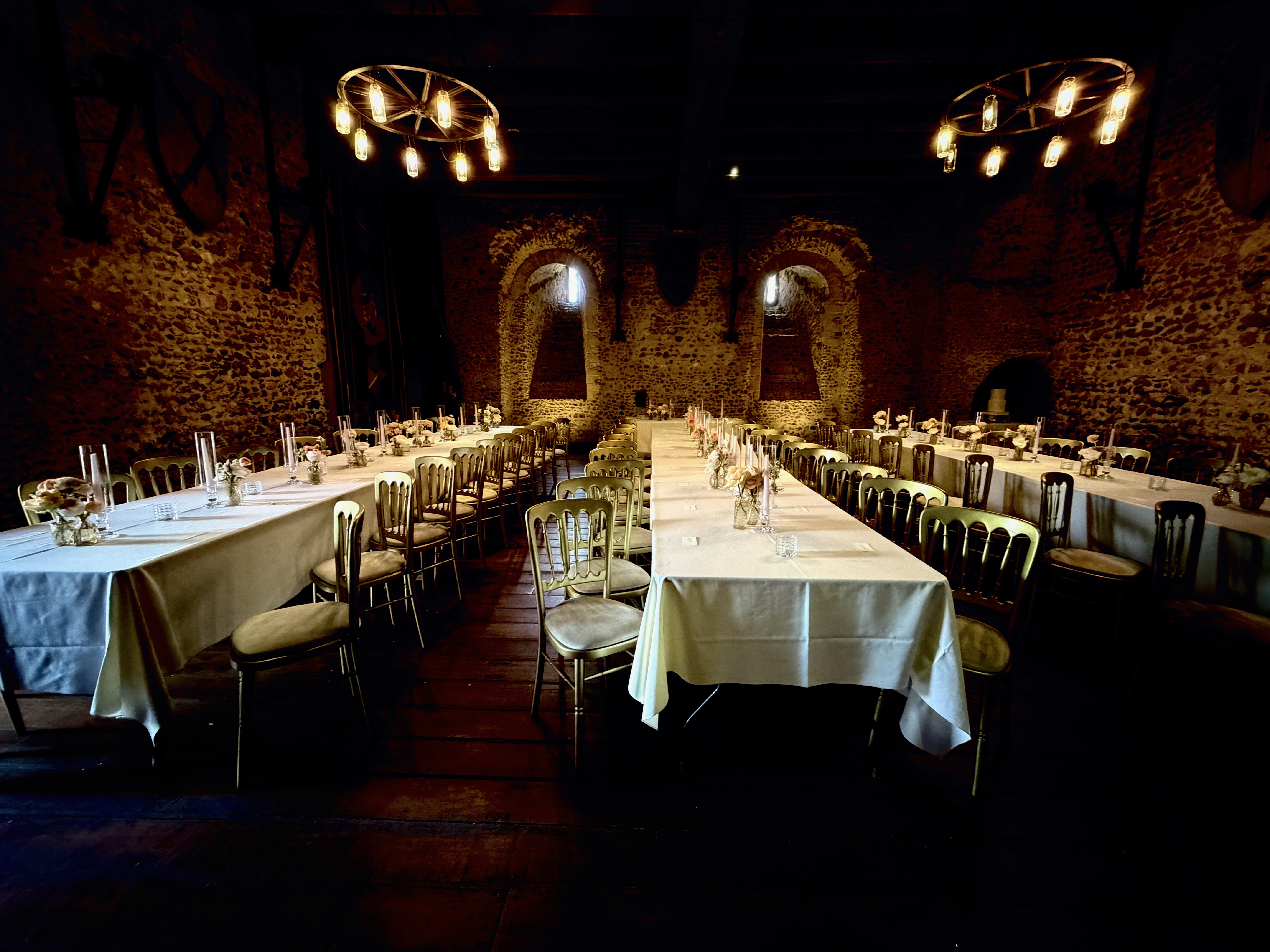 Long wedding reception tables with flowers and candles arranged across a stone-walled castle hall