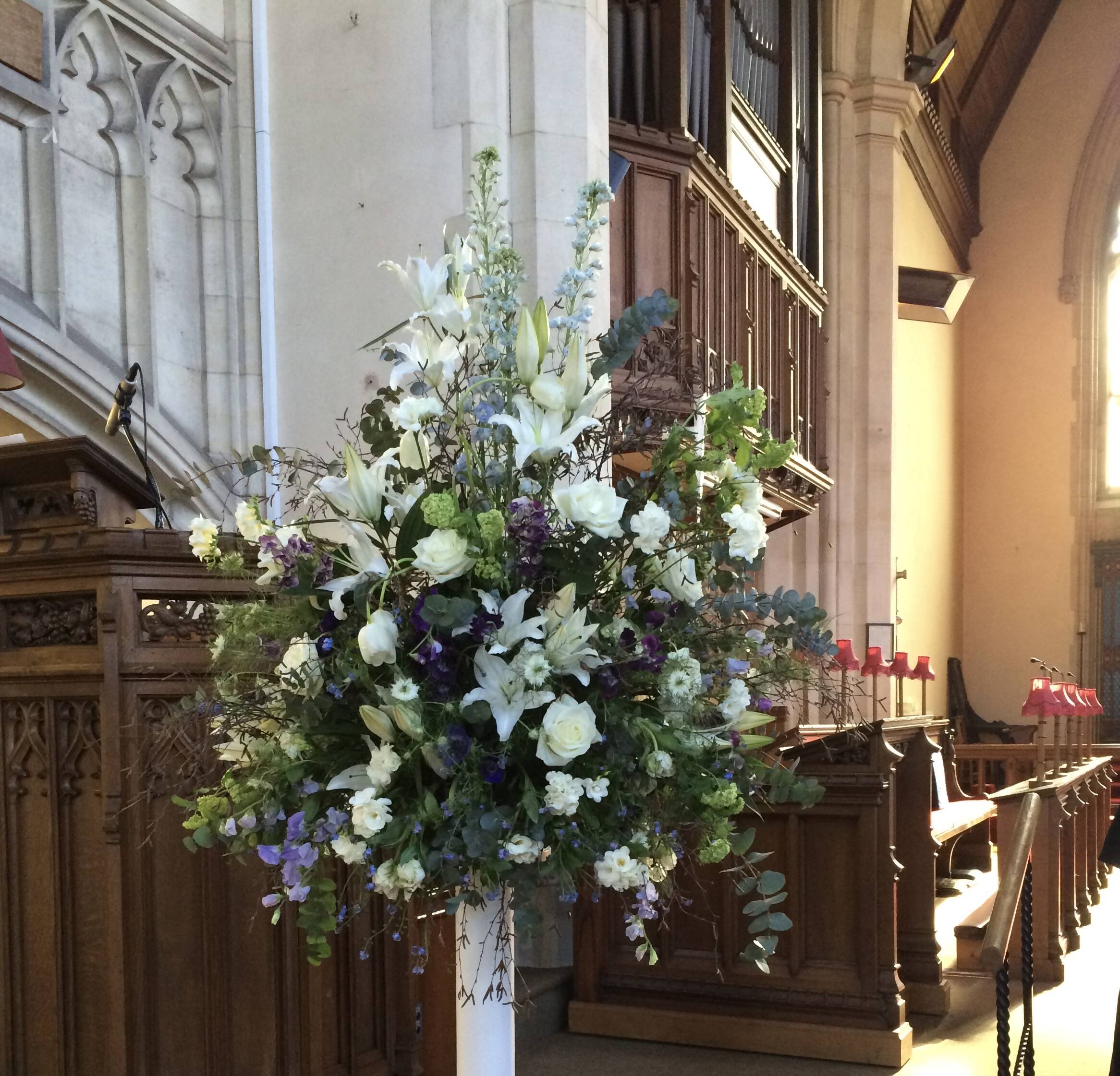 White lilies, pale blue flowers and foliage arranged beside the carved woodwork inside a church