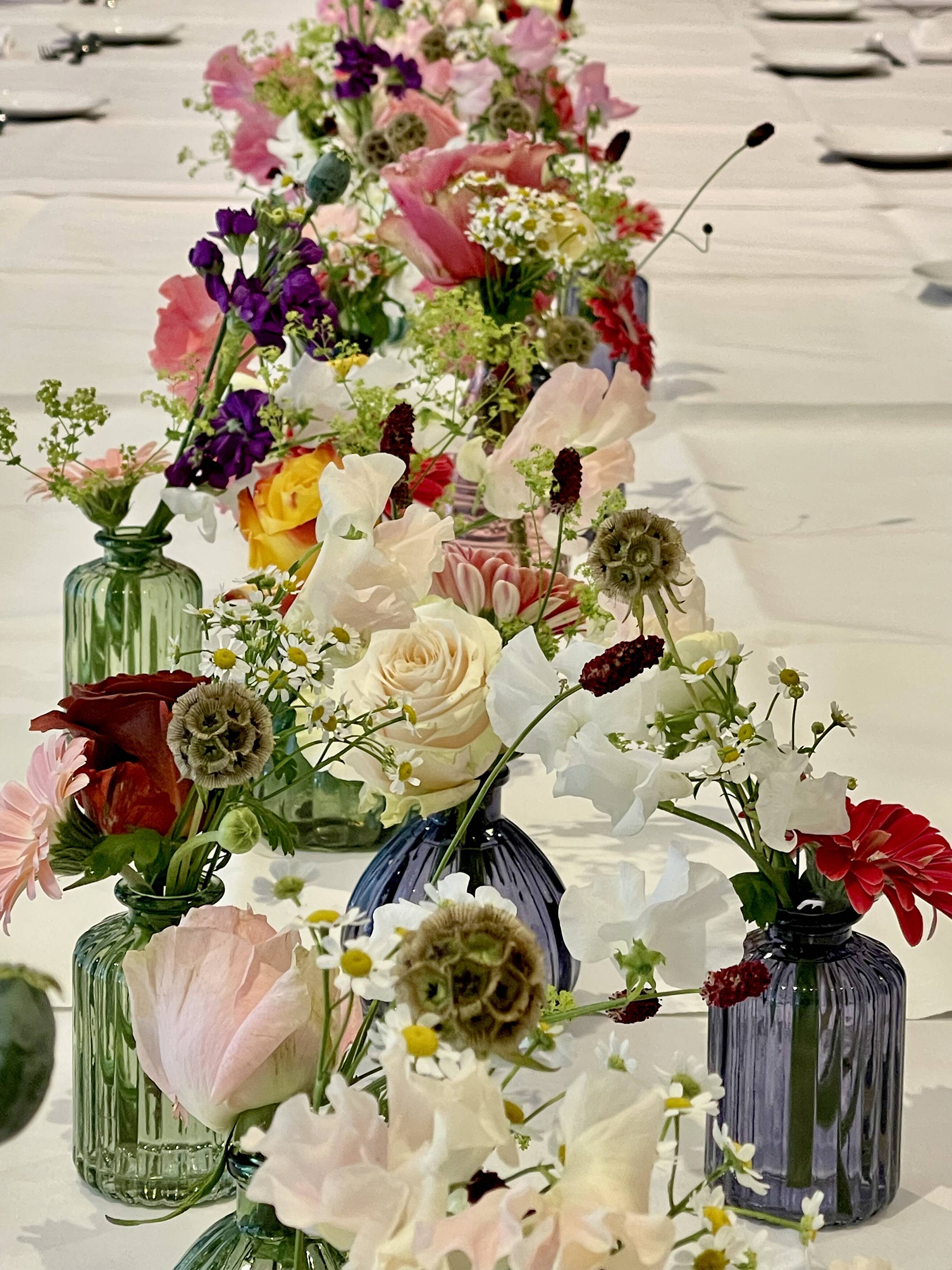 Individual glass vessels with colourful seasonal flowers arranged along a wedding table