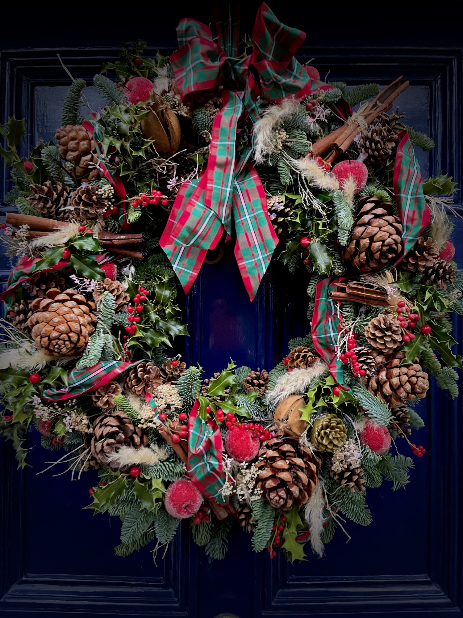 A generous Christmas wreath of evergreen foliage, pine cones, fruit and red tartan ribbon on a blue front door